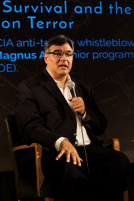 John Kiriakou, ancien officier de la CIA, assis sur une scène et tenant un micro lors d'une conférence publique